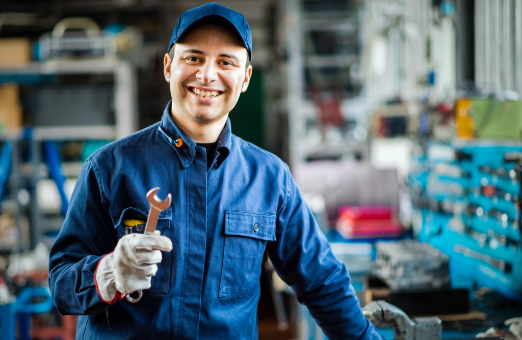 male mechanic in blue uniform smiling while holding wrench in his garage